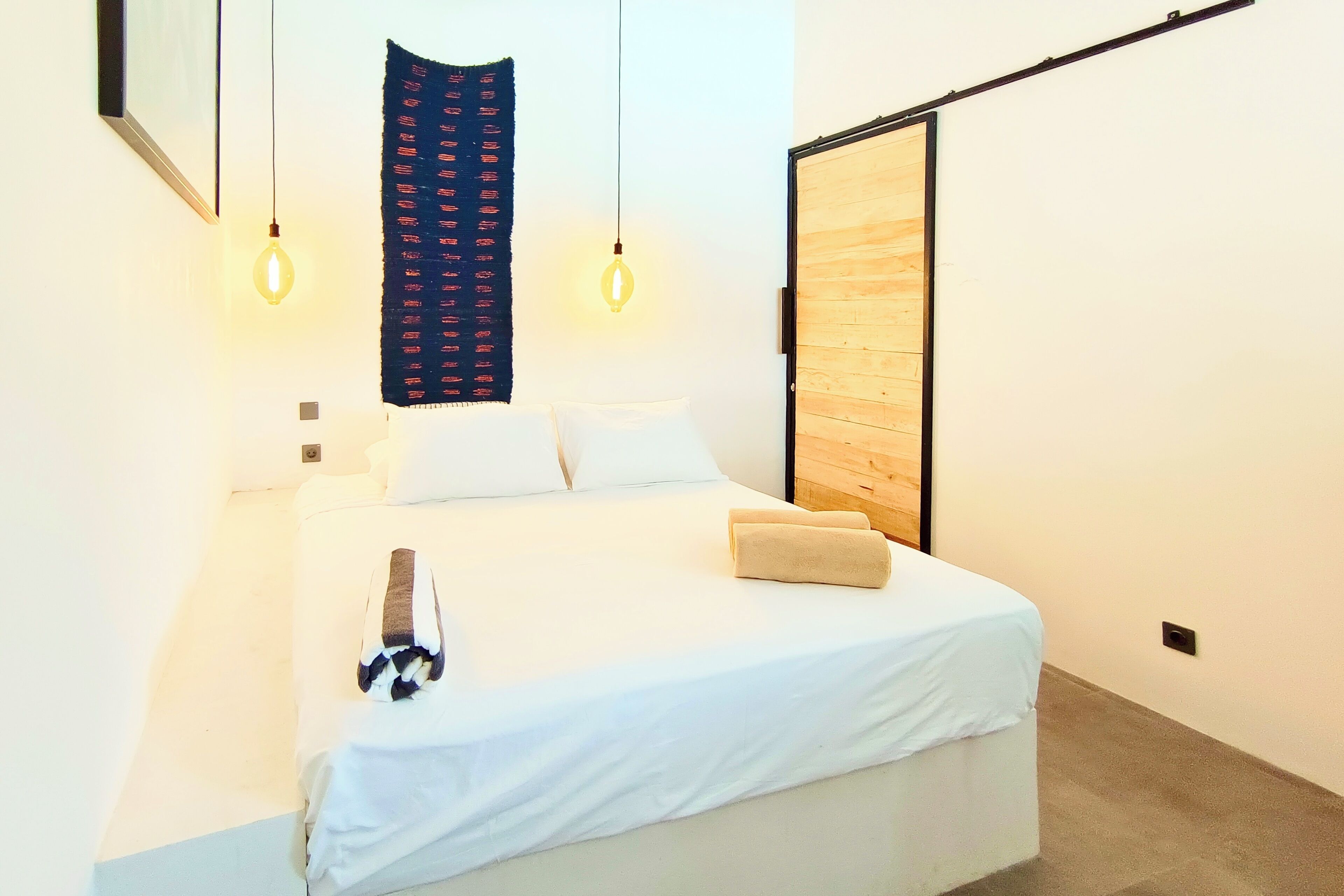 Un dormitorio moderno con una cama grande, dos lámparas colgantes, un panel de pared de madera y un tapiz oscuro con estampado.