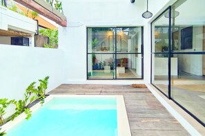 Un área de piscina moderna con terraza de madera, puertas de cristal que dan a un espacio habitable y plantas en macetas.