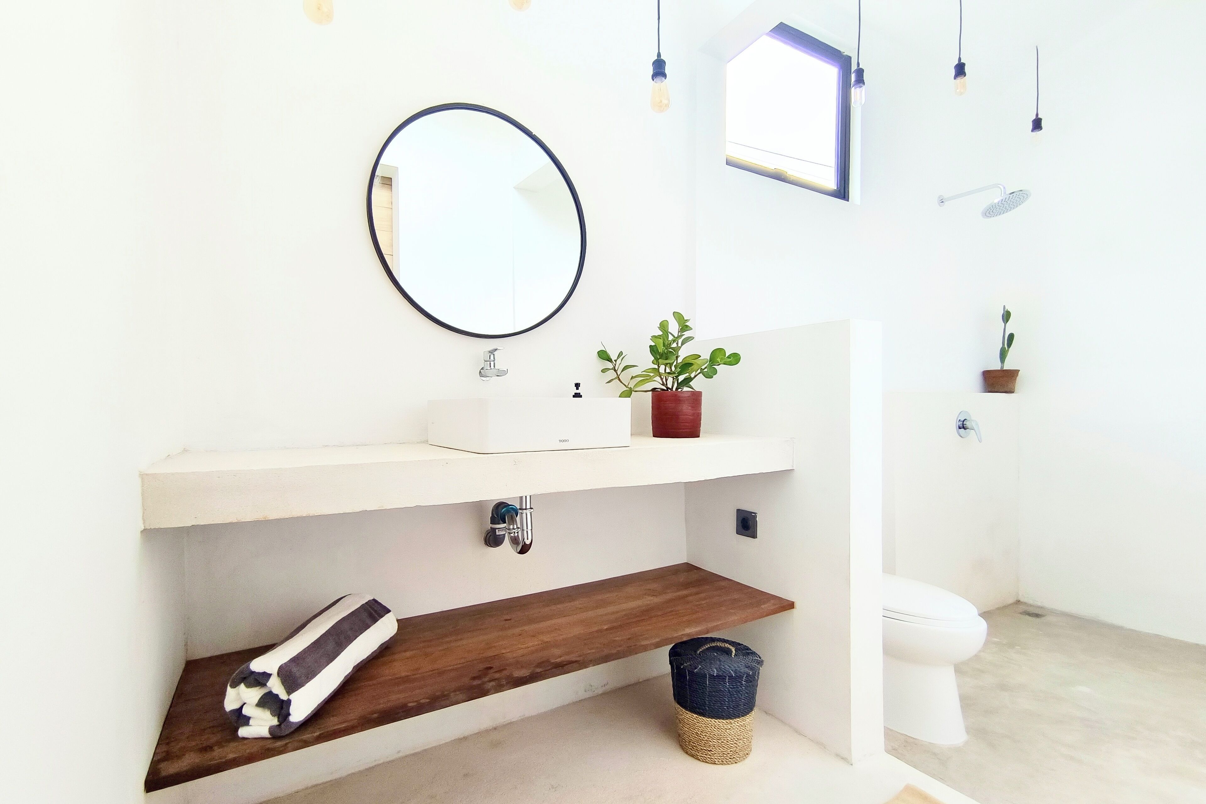 A bathroom with a white sink, a round mirror, a wooden shelf, a potted plant, a hanging light fixture, and a toilet.