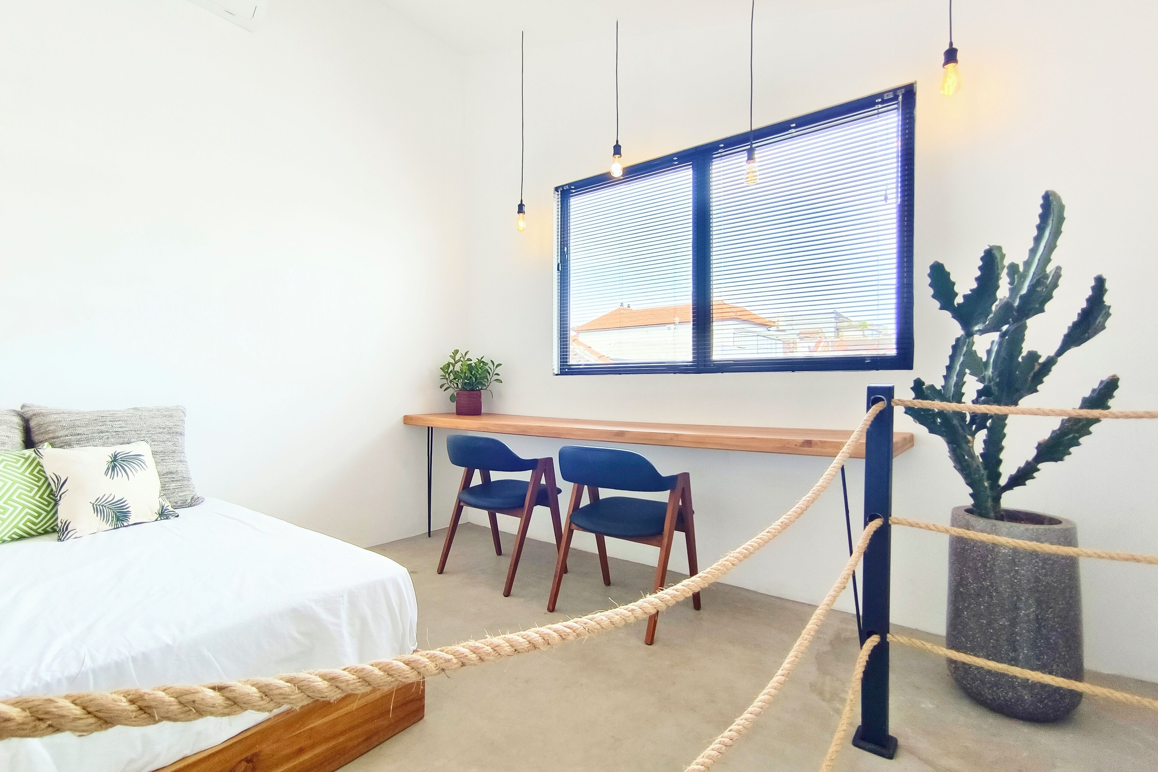 A bedroom with a bed, a desk, two chairs, a potted plant, and a window with blinds.