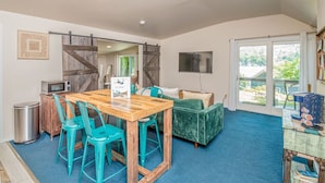 A cozy room with a wooden dining table, teal chairs, a green sofa, and a large window offering a view of greenery.