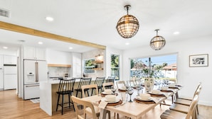 A modern kitchen with white cabinets, a central island, and a dining area with a wooden table set for six. - Designer Home - 9 Beds - New Pool - By Beach (Cocoa Beach)