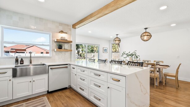 A modern kitchen with white cabinetry, a large island, and a dining area with wooden chairs and a table. - Designer Home - 9 Beds - New Pool - By Beach (Cocoa Beach)