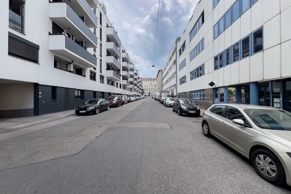 A modern urban street with multi-story residential buildings, parked cars, and a clear sky.
