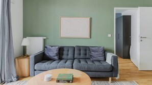 Una sala de estar con un sofá gris, una mesa redonda con libros, una lámpara y un cuadro en la pared.
