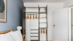 Un dormitorio con una cama, una mesita de noche, un armario con ropa colgada y una puerta.