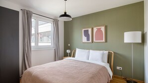 Ein Schlafzimmer mit einem Bett, einem Fenster mit Vorhängen, einer Stehlampe und zwei Wandbildern.