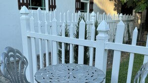 A patio with a round metal table and two chairs, a white picket fence, and a building with windows in the background. - The Tiny cottage in quite charming. Palos Verdes with AC, WiFi and washer/dryer  (Rancho Palos Verdes)