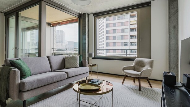 Un salon moderne avec un canapé gris, une table basse ronde et un fauteuil. De grandes fenêtres offrent une vue sur le paysage urbain.