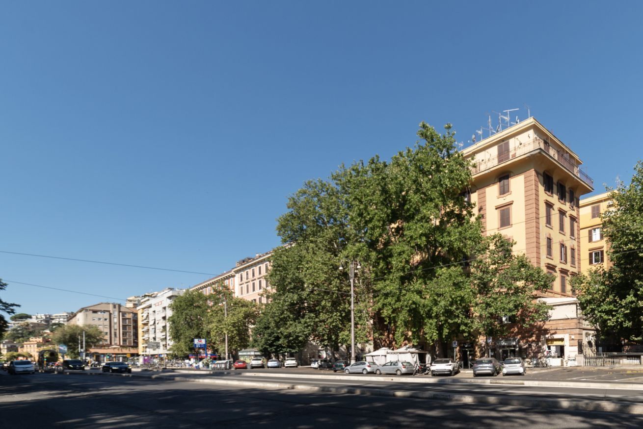 A street view with multi-story buildings, trees, and parked cars.