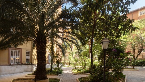 A paved courtyard with a palm tree, a lamp post, and a pathway leading to a building with barred windows. - GuestHost - Piazza Nievo in Trastevere Cozy Apartment (Roma)
