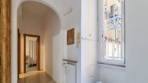 A hallway with a tiled floor, a radiator, a small table with a decorative item, and a window with a metal grille. - GuestHost - Piazza Nievo in Trastevere Cozy Apartment (Roma)