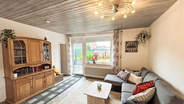 Ein gemütliches Wohnzimmer mit Holzbalkendecke, einer eingebauten Bücherreol und einem Sofa mit Kissen, einem Couchtisch und Blick ins Freie durch eine Glasdoor.