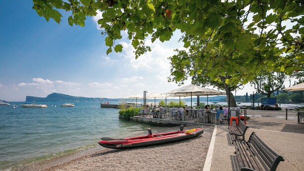 Un parc au bord du lac, avec une rangée de bancs, un canoë et un espace café doté de tables et de parasols.