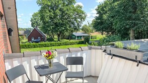 Eine Terrasse mit Tisch und zwei Stühlen, einem Blumenvasen und Blick auf eine grüne Landschaft mit Haus und Bäumen.