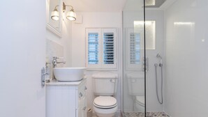 A bathroom with a white sink, toilet, and shower. There is a window with blinds and a mirror above the toilet. - Central Apartment with Patio - Sofa Bed (Cheltenham)