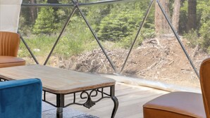 A room with a wooden table and chairs, a large window with a metal frame, and a view of a forested area.
