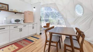 A compact kitchen with white cabinets, a microwave, and a pink appliance. There's a dining area with a wooden table and chairs, and a large window offering a view of trees.