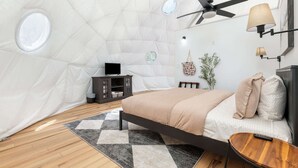 A cozy, well-lit bedroom with a bed, a small table, a ceiling fan, and a wall-mounted lamp.