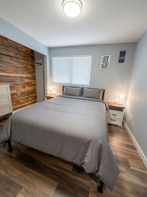 A bedroom with a bed, bedside tables, a wooden accent wall, a window with blinds, and a ceiling light. - Dog Friendly - Central AC - Light Rail to Seattle (Bothell)