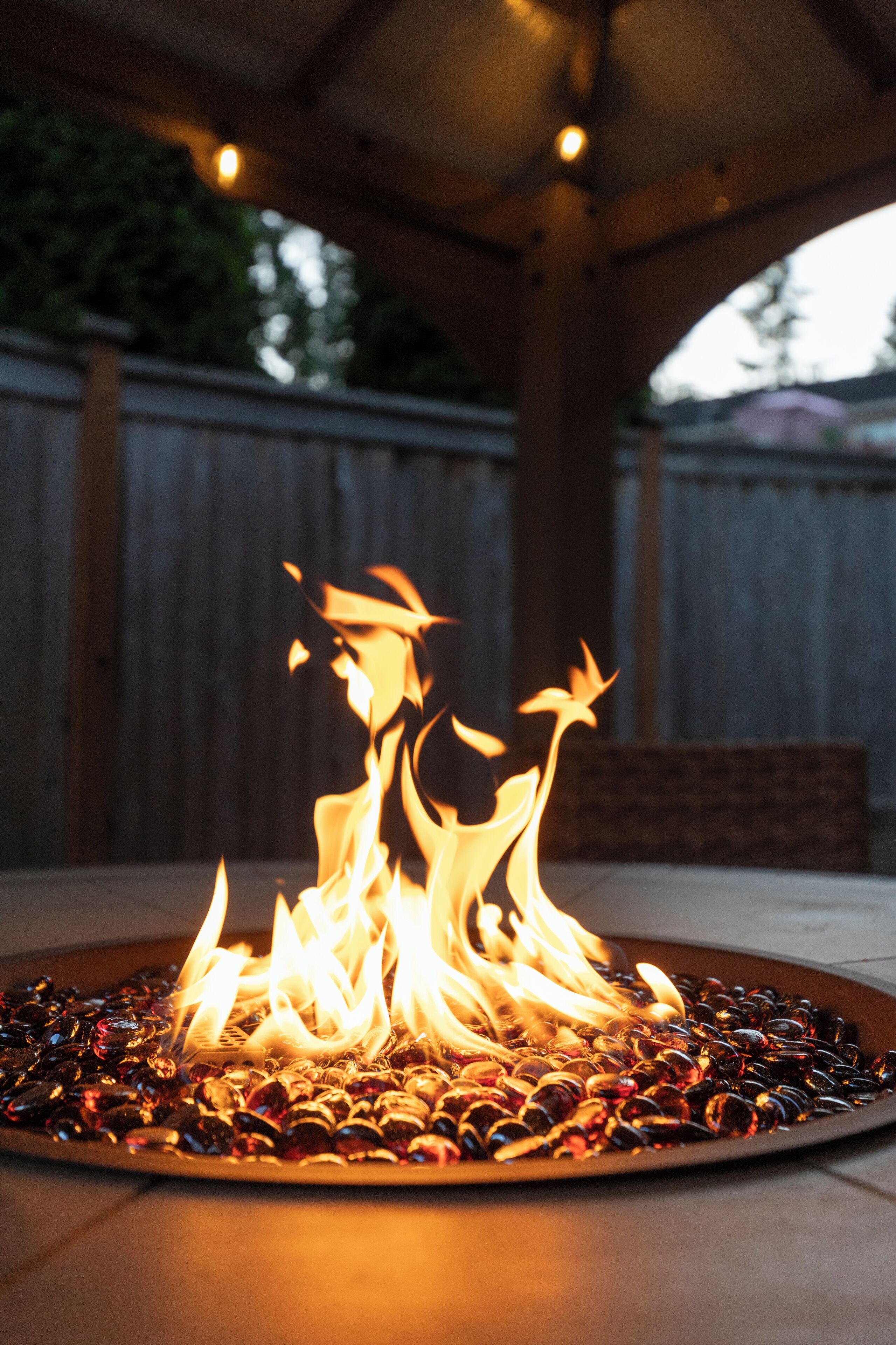 A fire pit with burning coals and flames, surrounded by a wooden deck and a wooden fence in the background.