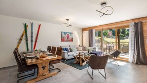 En rummelig opholdsstue med spisebord, sofa og stole, et billede på væggen og udsigt gennem glasdøren.