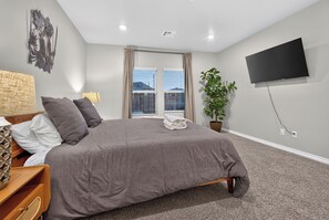 A bedroom with a bed, a TV, a plant, and a window with blinds.