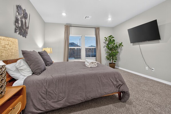 A bedroom with a bed, a TV, a plant, and a window with blinds.
