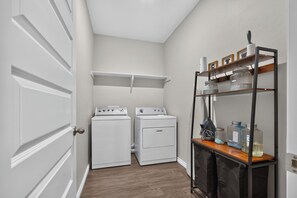 A laundry room with two white front-loading washers and dryers, a wooden shelf with various containers, and a door with a round handle.