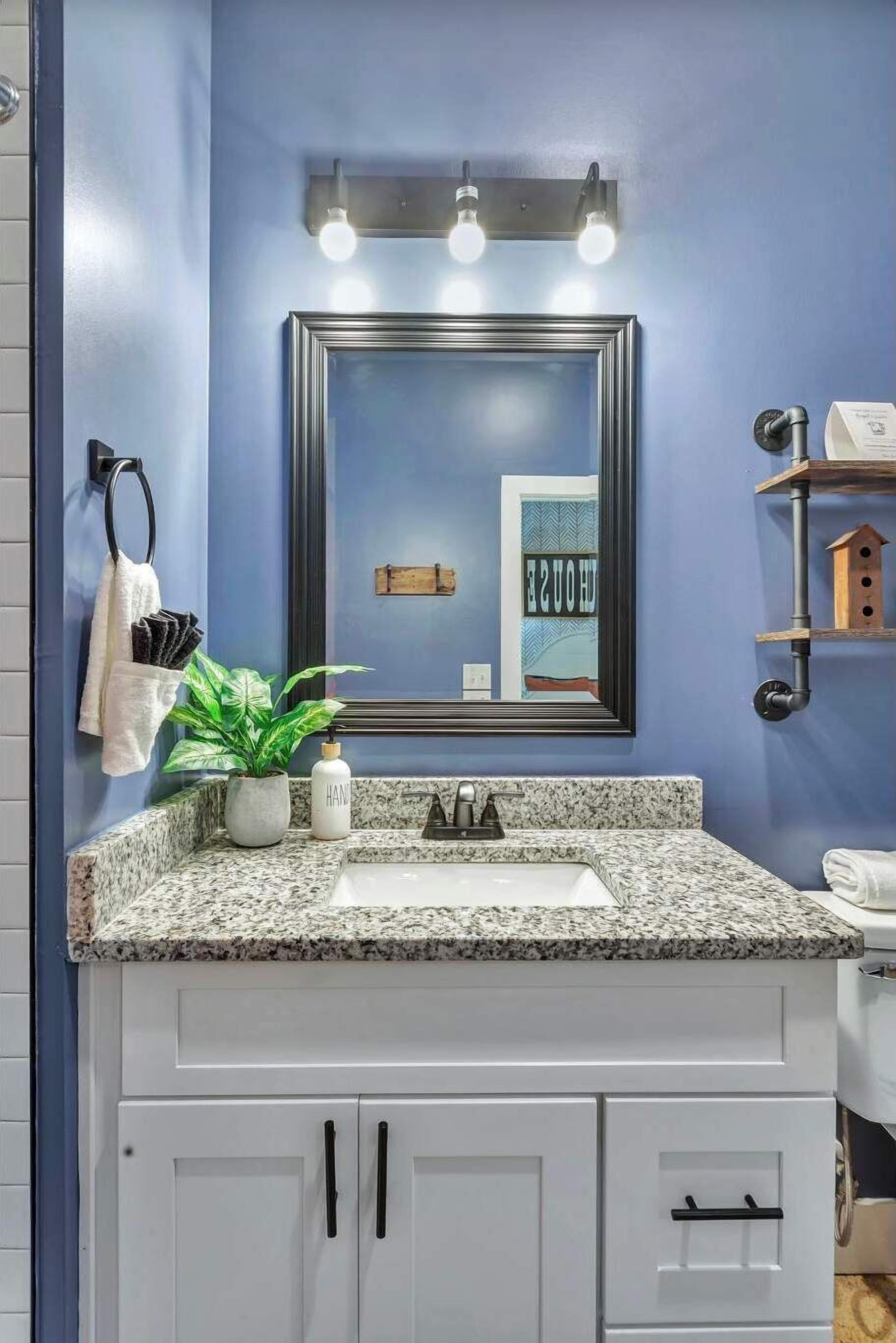 A bathroom with a blue wall, a granite countertop, a white sink, and a mirror with three lights.