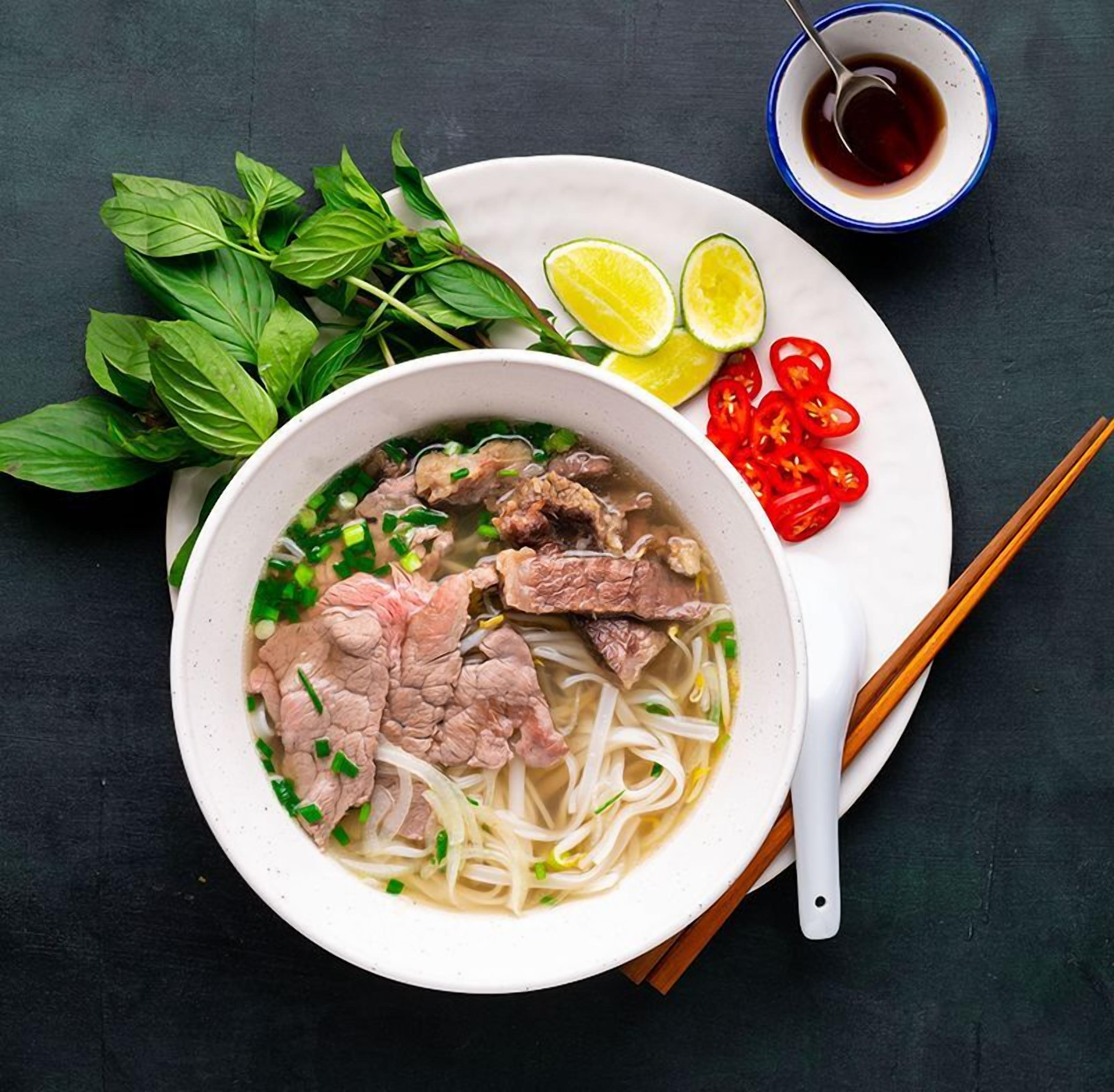 A bowl of noodle soup with beef, garnished with green onions and herbs, accompanied by lime wedges, sliced red chilies, and a small bowl of sauce on a dark surface.