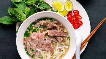 A bowl of noodle soup with beef, garnished with green onions and herbs, accompanied by lime wedges, sliced red chilies, and a small bowl of sauce on a dark surface.