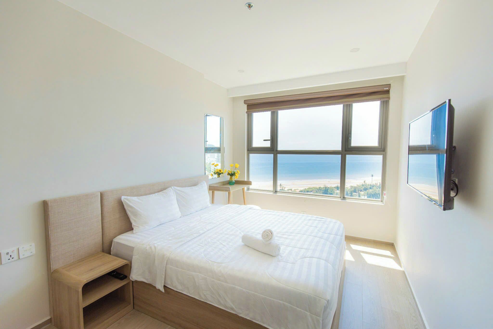 Quarto de hotel com uma cama grande, mesa de cabeceira, televisão na parede e janela com vista para a praia.