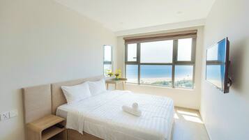 A hotel room with a large bed, a bedside table, a TV mounted on the wall, and a window with a view of the beach.