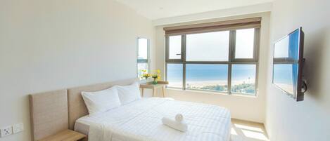 Quarto de hotel com uma cama grande, mesa de cabeceira, televisão na parede e janela com vista para a praia.