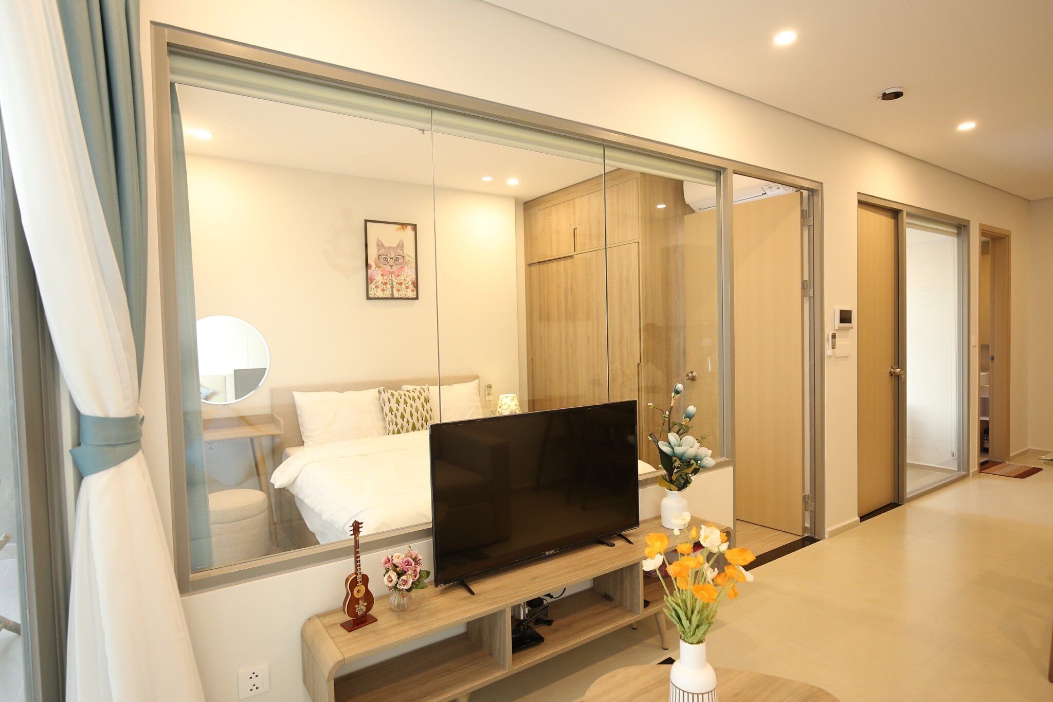 Quarto de hotel moderno com um espelho grande, televisão de tela plana, cama com roupa de cama branca, guarda-roupa de madeira e uma pequena mesa com um vaso de flores.