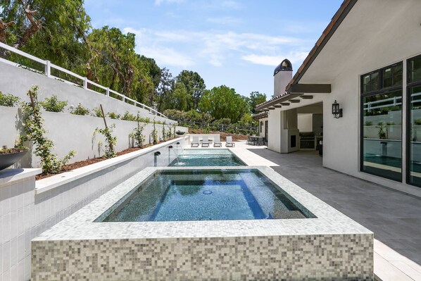Un área de piscina moderna al aire libre con cubierta de baldosas, zona de estar y un edificio con grandes ventanales.