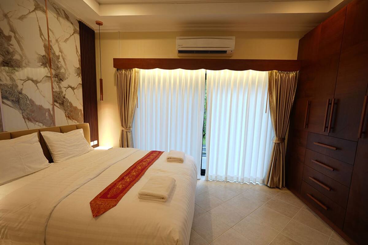 A hotel room with a large bed, a wooden wardrobe, and a view through sheer curtains.