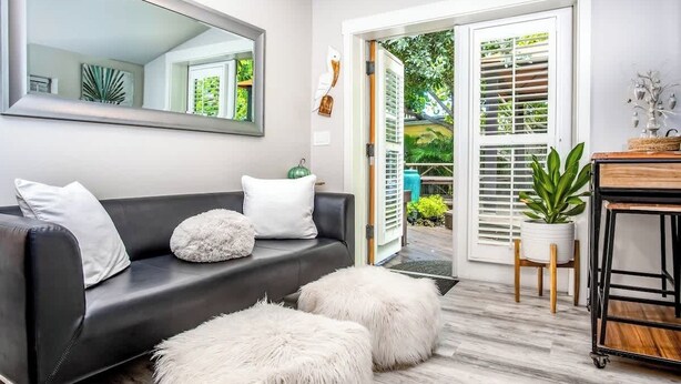 A modern living room with a black leather sofa, white fluffy ottomans, a large mirror, and a wooden coffee table. - Honeymoon Banyan Suite near Higgs Beach (Key West)