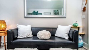 A modern living room with a black leather sofa, white cushions, and a fluffy round ottoman. A large mirror hangs above the sofa, reflecting the room. - Honeymoon Banyan Suite near Higgs Beach (Key West)