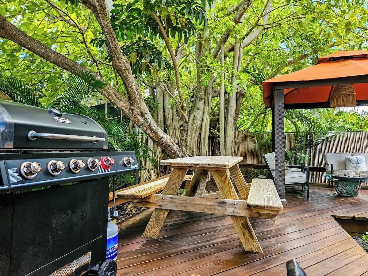 Une terrasse en bois équipée d’un barbecue, d’une table de pique-nique et d’un espace salon extérieur, le tout entouré d’une végétation luxuriante.