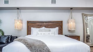 A large bed with a wooden headboard, two bedside lamps, a fluffy throw blanket, and a pillow with the text "ALL ARE WELCOME HERE".