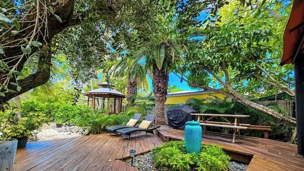 A wooden deck with lounge chairs, a gazebo, and a barbecue area surrounded by lush greenery.