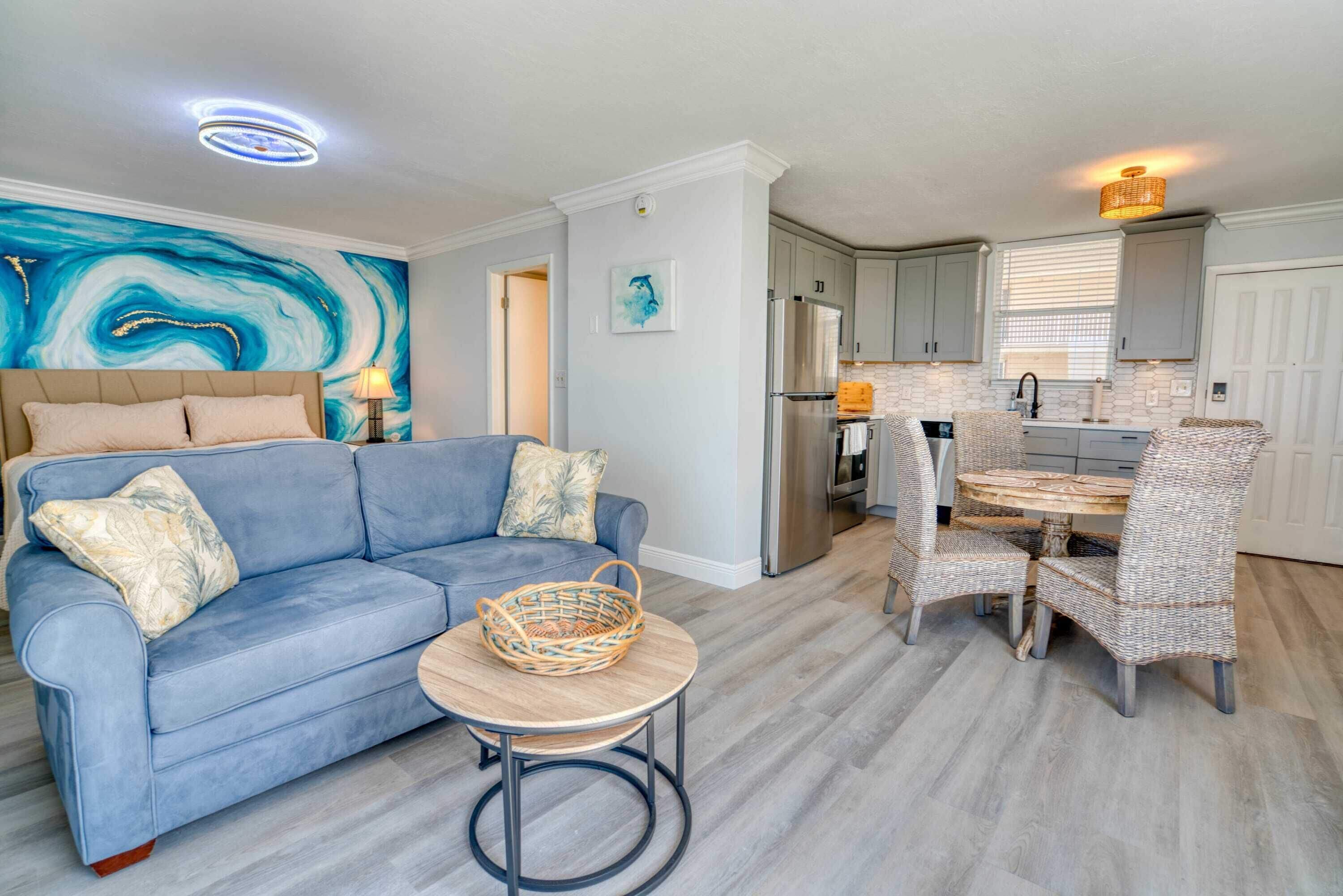 A living room with a blue sofa, a round table, wicker chairs, and a kitchen area with white cabinets and a refrigerator.