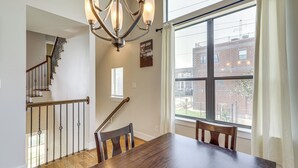 Un espace repas avec une table et des chaises en bois, un lustre, un escalier et une grande fenêtre donnant sur les bâtiments environnants.