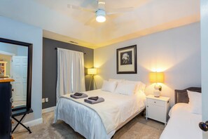 A hotel room with a bed, bedside lamps, a ceiling fan, a framed picture on the wall, and a door leading to another room. - Heated Pool, Hot Tub & Fire Pit in Ruby Park (Eugene)