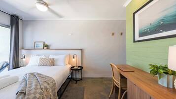 A hotel room with a bed, bedside table, lamp, chair, desk, and a large framed picture on the wall.