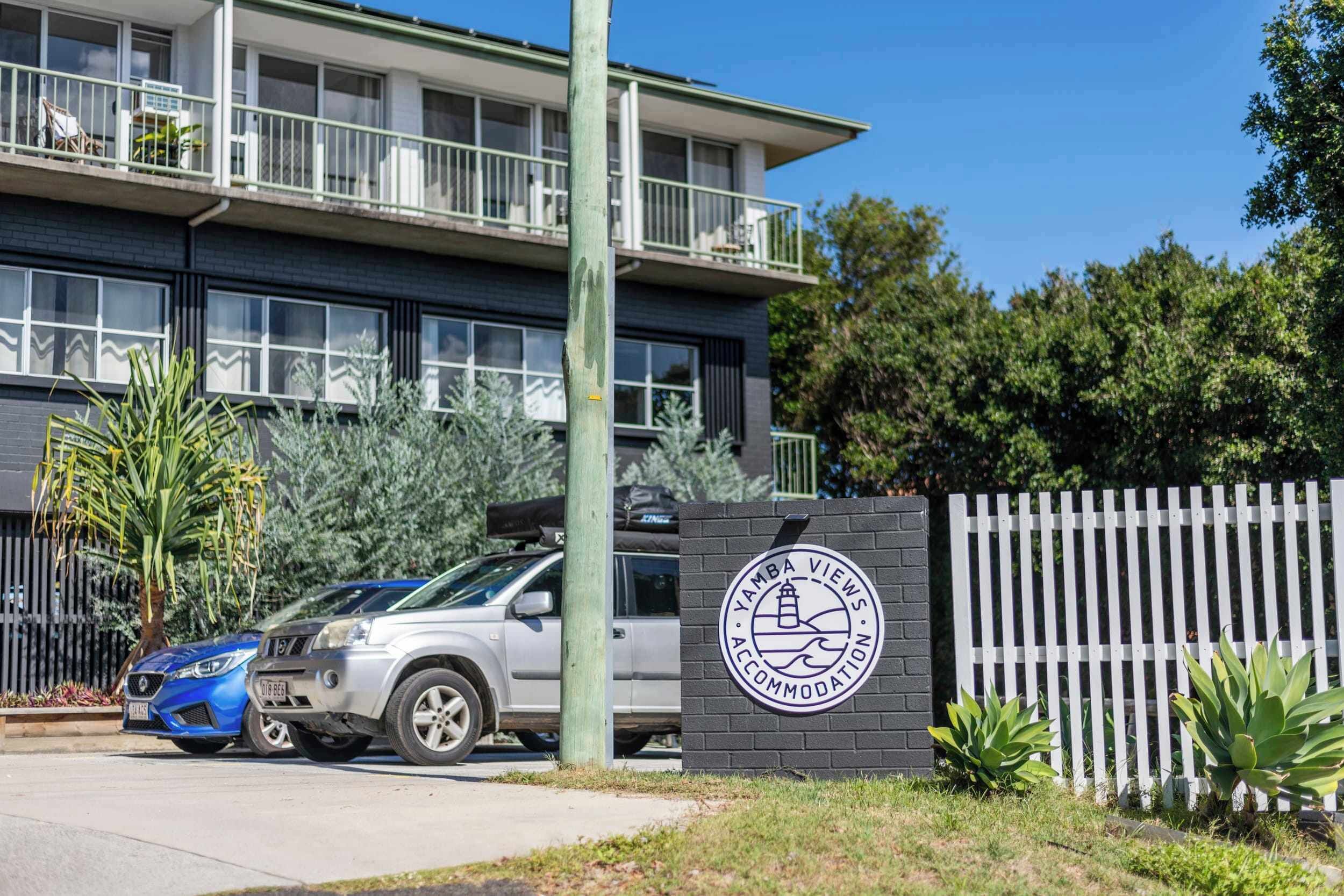 A multi-story building with balconies, a parking area with two cars, and a sign indicating 'YAMBA VIEWS ACCOMMODATION.'