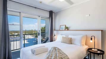 A hotel room with a large bed, a bedside table with a lamp, a view of the city through large windows, and a balcony.