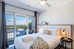 Habitación de hotel con una cama grande, una mesita de noche con lámpara, vistas de la ciudad a través de amplias ventanas y un balcón.